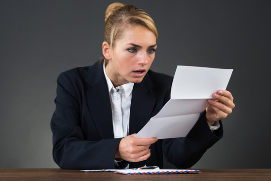 Shocked young businesswoman reading letter at desk over gray background